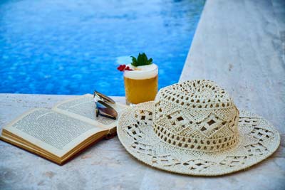 A photo of an open book with a pair of sunglasses on top of it, a sun hat, and a cocktail drink, all put together by the pool