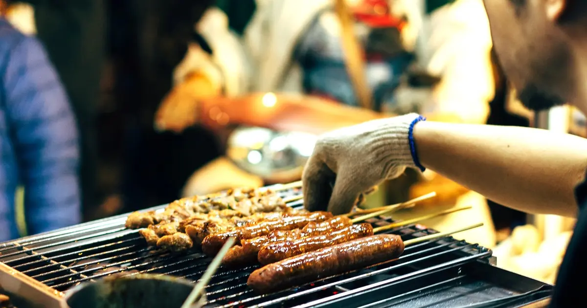 A man grilling pork barbeque and hotdogs on a stick, which are popular finger food pulutan.